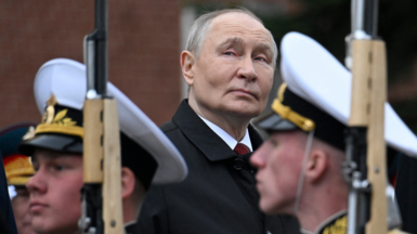 Vladimir Putin stands at the centre of a ceremonial honour guard, surrounded by uniformed service members in full dress. The scene is outdoors, framed by brick architecture and greenery. The military personnel wear white caps, dark uniforms with gold insignia, and carry rifles held in precise ceremonial positions. Their posture is rigid and disciplined, forming a structured corridor around Putin. Vladimir Putin is dressed in a dark overcoat and red t. He appears to be observing or participating in an inspection.
