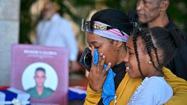 A woman wipes her tears while hugging a young girl, as a picture of a soldier can be seen in the background