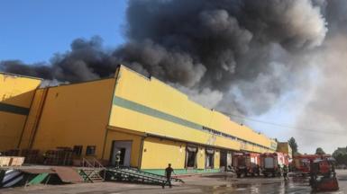 A thick, black cloud of smoke hovers above a yellow building. Fire trucks are parked in front