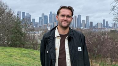 A young white man with brown hair, dressed in brown tshirt, white shirt and black jacket smiles at the camera with a backdrop of trees and skyscrapers