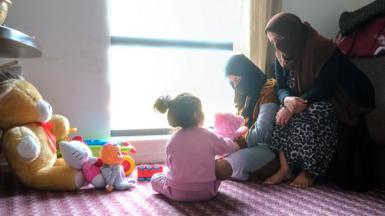 Photo of a hotel room with three people - a women and older child with their faces covered, and a younger girl with a back to the camera sitting on a patterned pink carpet