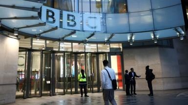 A person wearing a white shirt and carrying a shoulder bag walks towards the entrance doors at BBC Broadcasting House in London on Monday, with the BBC logo in view.