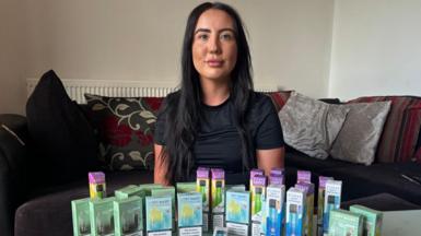 Cara is a 25-year-old woman with long dark hair, she is wearing a black T-shirt and she is sitting on a sofa. On a table in front of her is an array of multi-coloured disposable vape boxes.