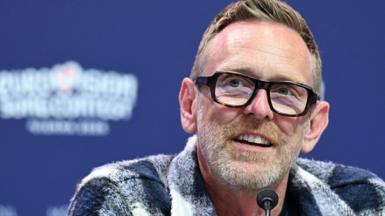 Martin Green wearing glasses and a grey and white checked shirt, seated and speaking into a small microphone in front of a blue backdrop with the Eurovision logo