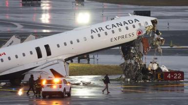 Air Canada flight AC8646 with its nose upturned and wrecked. Emergency services surround it. The tarmac is wet, it is early morning. 