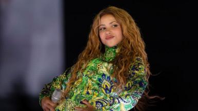 Shakira in a green top, her long brown hair loose, smiling on stage