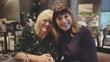 Two women, one with blonde hair and one with brown hair, hold hands and smile at the camera.