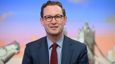 Darren Jones face and shoulders. He has brown wavy hair and is wearing circular glasses with a blue suit, blue shirt, and red wool tie. Behind him, in soft focus, is some artwork of UK landmarks, including London's Tower Bridge