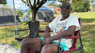 A man in a white t-shirt and aqua shorts and black sunglasses sits in a camping chair on a patch of grass. A second camping chair is next to him, and tents and a wheelchair are visible in the background. In the far distance, a large building is visible.