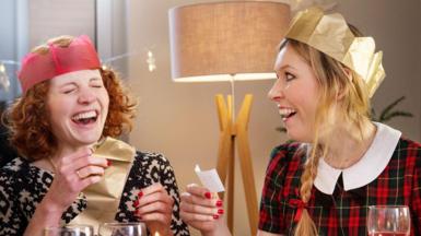 Two women wearing Christmas hats are sharing a joke from a pulled cracker.