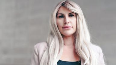 A head and shoulders portrait of Rebecca, standing against a light stone background. She has long blonde wavey hair and wears a black top with a light pink jacket. She looks pensively into the camera. 