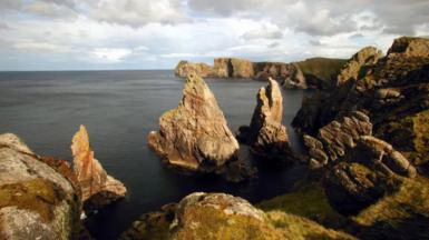 A stock image of Tory Island coastline - there are rocks emerging from the water, there is grass on the coastline.