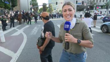 Lauren Tomasi, who wears casual clothes and has blonde tied back hair, holds a mic as she speaks to the camera few metres from a police line