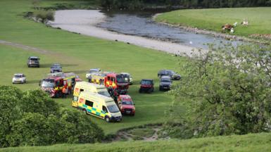 Ambulances, fire engines and other vehicles parked on a trimmed field close to a river meandering nearby.