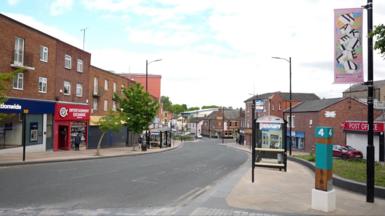 A row of shops with a road in the centre. A bus stop is on one of the footpaths and there are artistic pink signs reading Wakefield attached to the lamp posts.