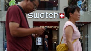 People walk past a store of Swiss watchmaker Swatch, in Beijing, China. A woman and a man can be seen in the foreground. He is wearing a maroon t-shirt and she is wearing a pink top with a yellow bag. The Swatch logo can be seen shining behind them, with the entrance to the store below