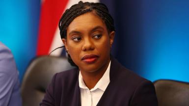 Conservative Party Leader Kemi Badenoch seated in front of a blue backdrop with part of the Union Jack flag visible. She is wearing a dark blazer over a white shirt, with braided hair