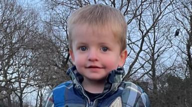 Two-year-old Alfie standing wearing a check-print blue coat in park
