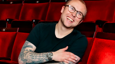 Joe in a black top, wearing glasses, sitting in red theatre/cinema seats