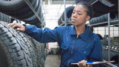 A female car mechanic wearing blue overalls stands with her hand on a car wheel in a wheel warehouse.