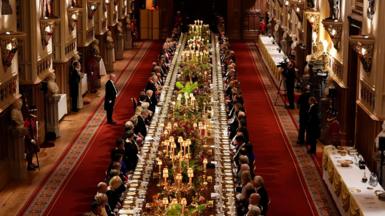A long dining table with dignitaries seated down either side is seen in a banquet hall, with staff and press against the walls.