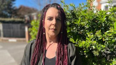 Sonia is staring directly at the camera. She has braided hair that has a slightly red tint. She is wearing a dark shirt and sitting on the sofa in her lounge. She looks thoughtful.