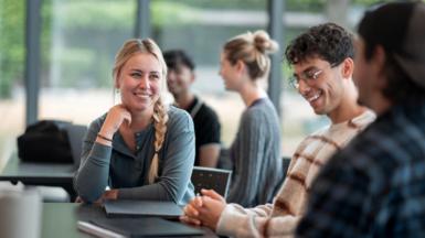 A stock image shows four young people seated at a table in a bright, modern classroom or seminar room with large windows. They are engaged in conversation with notebooks and folders on the table.