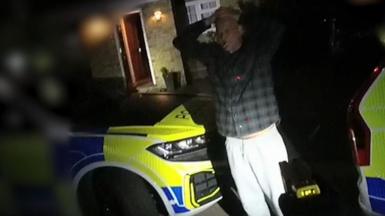 Jeremy Heath has his hands on his head, wearing a dark-coloured check shirt, and standing outside a house in darkness. There are two marked police cars parked either side of him. He appears to have a cigarette in his mouth.