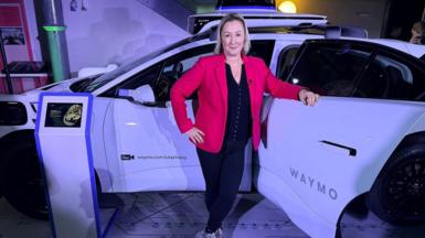 BBC technology editor Zoe Kleinman stands in front of a white car, wearing a red jacket over a black top and black trousers 