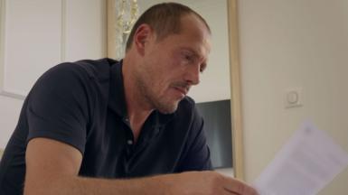 A man with short, light brown hair and stubble looks at a piece of paper. He is wearing a dark blue, short-sleeved polo shirt. 