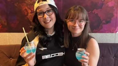 Caitlin and Katy sit on a purple sofa smiling while holding light blue BuzzBall canned cocktail drinks.