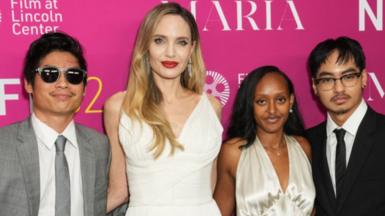 An image of Angelina Jolie wearing a white dress and three of her children on the red carpet standing in front of a pink board 