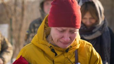 Natalia stands weeping by her husband's grave in a snowy cemetery. She's wearing a red beanie and a yellow jacket, and holding a red rose