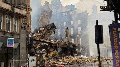 The Union Corner building is reduced to rubble after a fire in Glasgow. Victorian buildings and street furniture surround the iron and sandstone remains. 