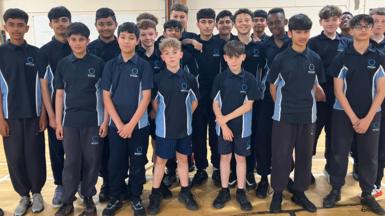 A group of Year 8 boys stand shoulder to shoulder dressed in navy and blue school PE kit for a PE lesson on an inside basket ballcourt