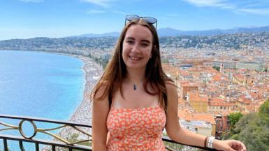 Claire wearing an orange sundress overlooking the coastline of Nice, France. There are orange buildings in the background the sea on the right.