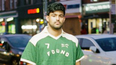 Sufiyaan Salam wearing a green and white T-shirt with the Found logo, standing at dusk in front of cars and restaurants on the Cutty Mile