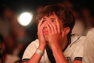 A man wearing an England shirt partially covers his face with his hands and looks close to tears.