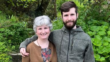 Anne and Will Thompson stand in a garden with trees behind them. He has his arm around her shoulder and both are smiling.