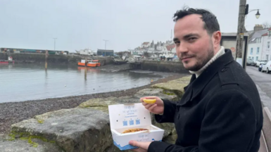 James Muirhead na sheasamh aig balla-mara ann am baile ann am Fìobha a' cumail suipear èisg is tiops aige 