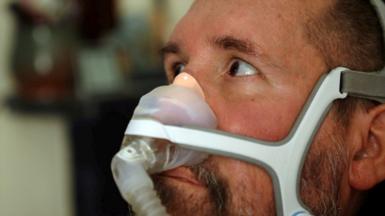 A close up of Nik Ward, an MND sufferer, sitting in his motorised wheelchair while he looks towards a family member standing to the left of the camera. He has a breathing apparatus across his nose.