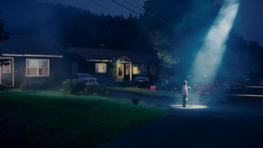 A person stands in a driveway in front of a home as a spotlight shines on them