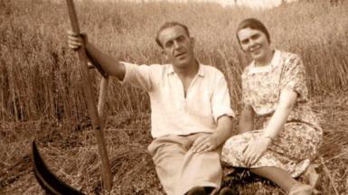 Fear le speal na làimh 's e na shuidhe air dlòthan coirce air am buain 's boireannach na suidhe ri thaobh. Coirce fhathast ri bhuain air an cùlaibh.