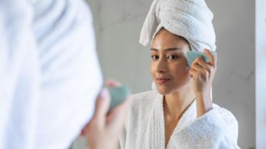 Woman wearing bathrobe and towel using a gua sha stone on her face, looking at herself in the mirror