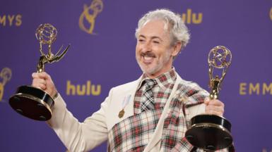 Alan Cumming celebrates the Traitors winning two Emmys in 2024 - he is grinning broadly while holding two Emmy awards up, one in each hand. He is wearing a suit with a tartan plaid design.