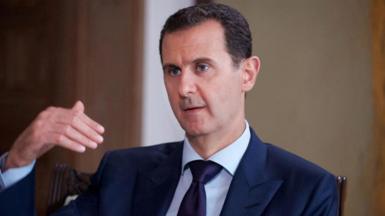 Bashar al-Assad pictured in 2016 speaking while gesturing with one hand. He is wearing a dark suit, white shirt and tie.