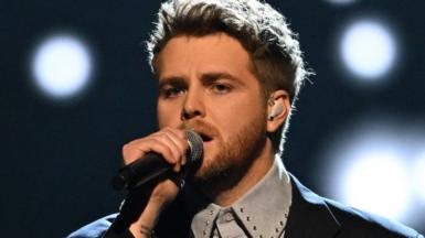 Singer-songwriter Alex Warren holds his microphone as he performs at the American Music Awards. Stage lights shine behind him.