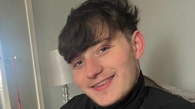 John Mcnab smiles at the camera while sitting on a couch in a living room. He has short, dark brown hair and is wearing a dark top.