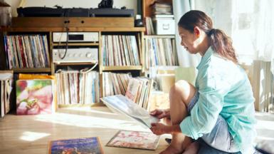 A woman kneels on the floor surrounded by music albums in a room with shelves filled with record albums.