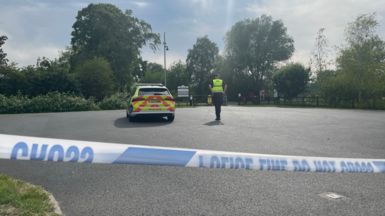 A police officer next to a police car in a car park with police cordon tape across the image